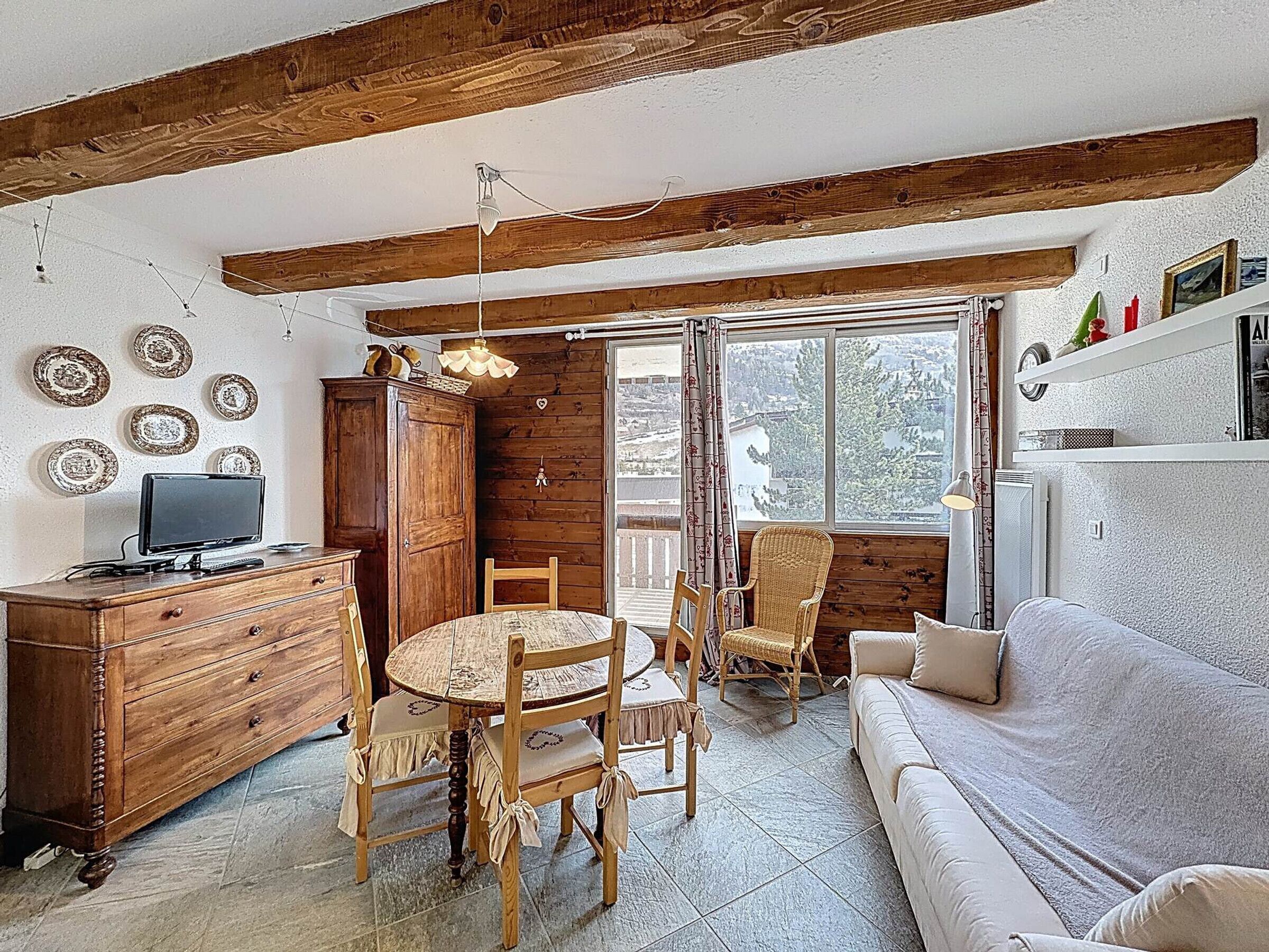 A cozy room with a wooden ceiling, a dining table with chairs, a sofa, a television, and a window with a view.