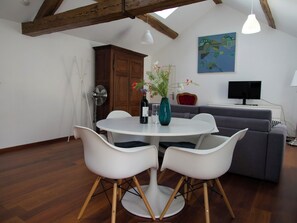 Un coin repas avec une table ronde, des chaises blanches, un vase de fleurs et un téléviseur en arrière-plan.