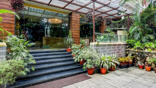 Entrada de hotel con escalones que conducen a una puerta de vidrio, rodeada de plantas en macetas y un área techada con barandilla de metal.