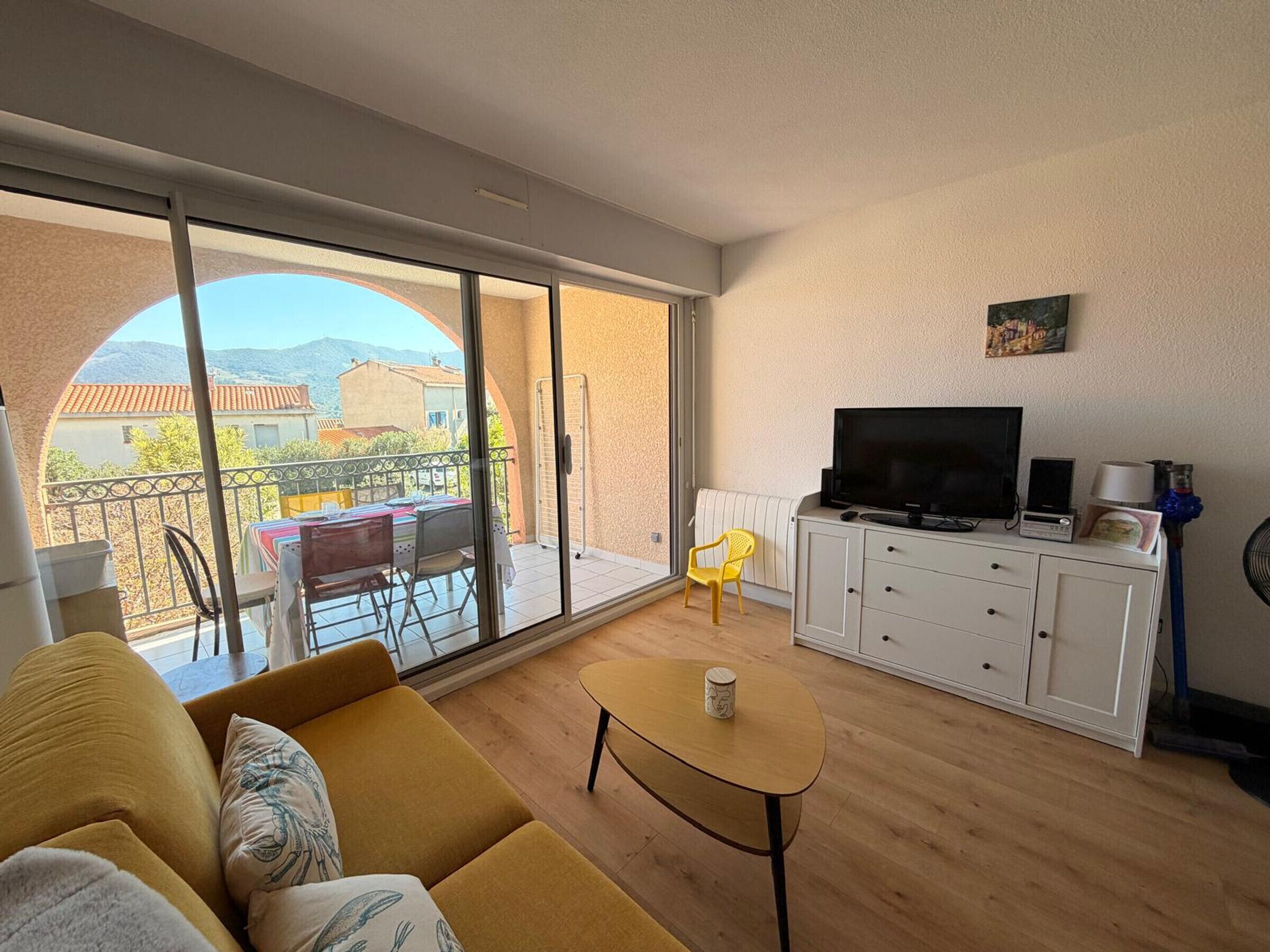 A living room with a sofa, a coffee table, a TV on a cabinet, and a balcony with a table and chairs.