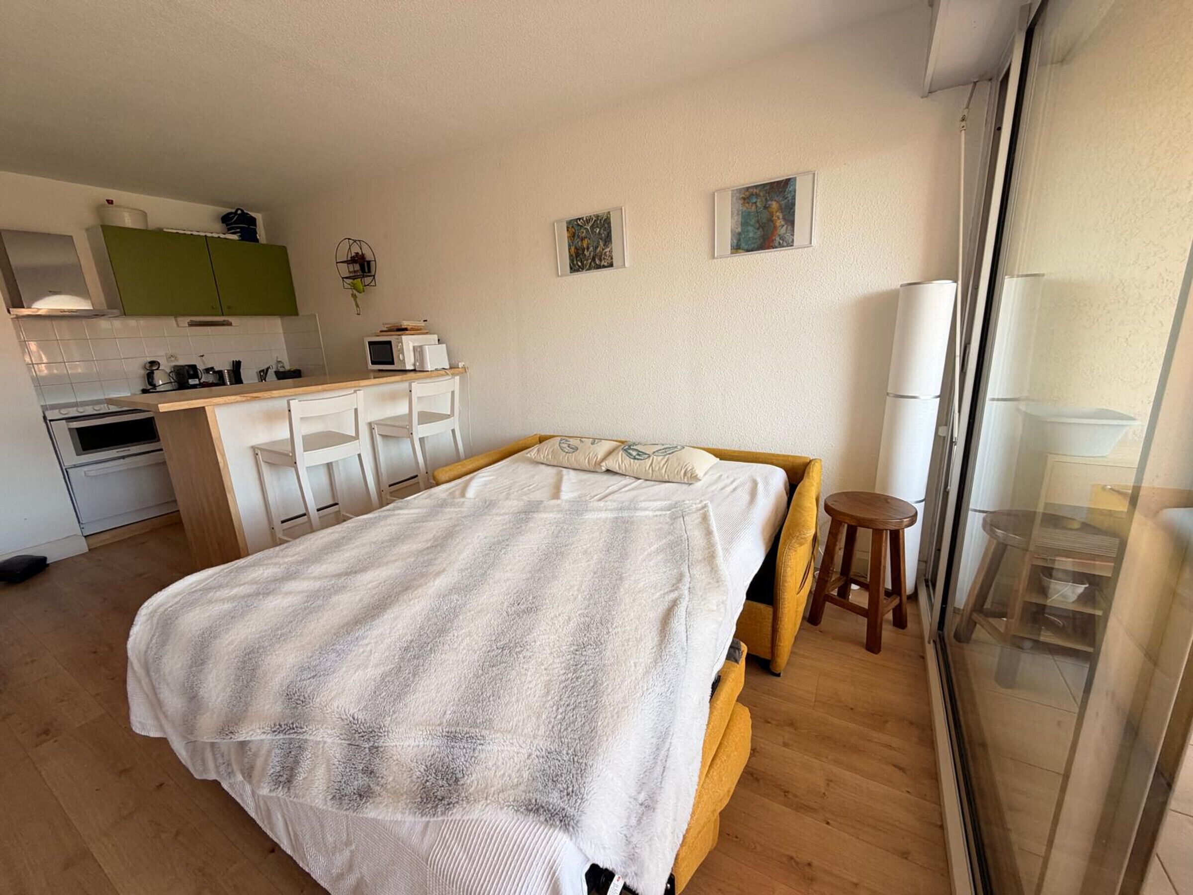 A bedroom with a bed, a small stool, a window with a view, and a kitchenette in the background.