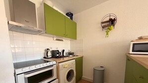 Ett kompakt kök med en spis, ugn, tvättmaskin, mikrovågsugn och en liten hylla med diverse föremål.