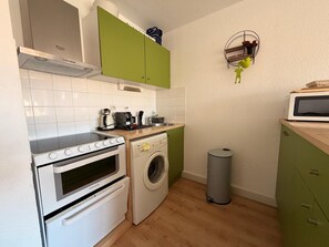A compact kitchen with a stove, oven, washing machine, microwave, and a small shelf with items. - Apartment with parking, mountain view - 4 guests (Banyuls-sur-Mer)