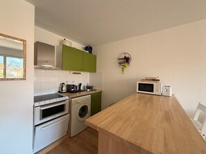 A compact kitchen with a washing machine, oven, microwave, and a wooden table. - Apartment with parking, mountain view - 4 guests (Banyuls-sur-Mer)
