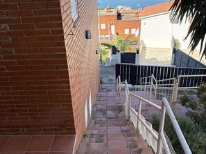 Eine schmale Backstein-Treppe mit Metallgeländer, die zu einem befestigten Bereich führt, mit Blick auf das Meer und Gebäude im Hintergrund.