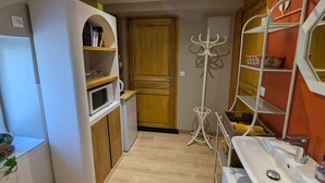 Een kleine keuken met een magnetron, koelkast, houten deur, kapstok en een wastafel met spiegel.