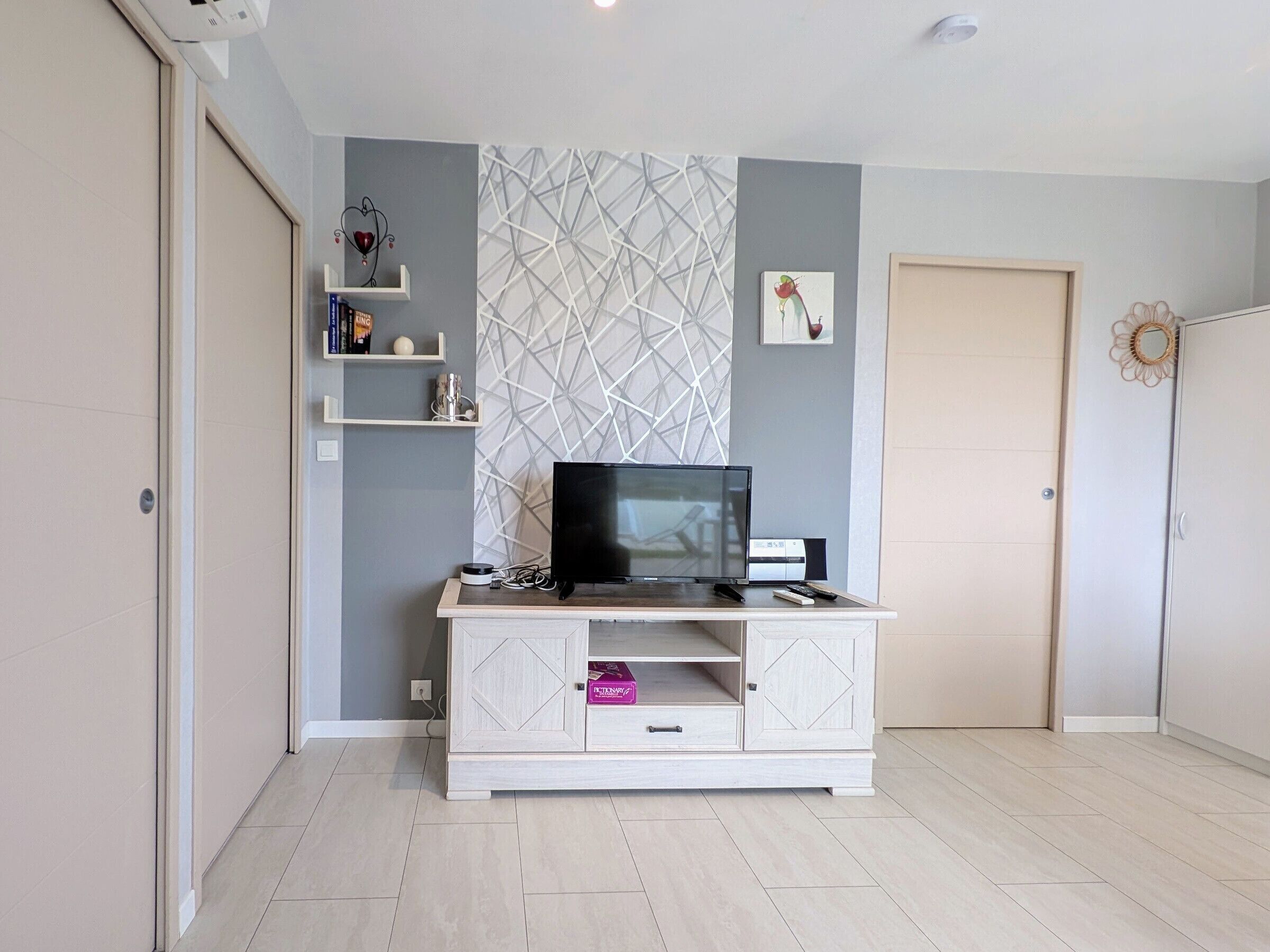 A room with a TV on a white cabinet, a geometric wall pattern, a door, and a shelf with books and decorative items.