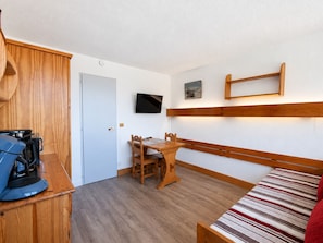 Ein kleines Zimmer mit einem Bett, einem Schreibtisch und einem Stuhl. Ein Fernseher ist an der Wand montiert und eine Tür führt in einen weiteren Raum.