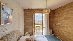 Une chambre à coucher avec un lit, un luminaire suspendu, un tableau au mur et une vue sur l’extérieur.