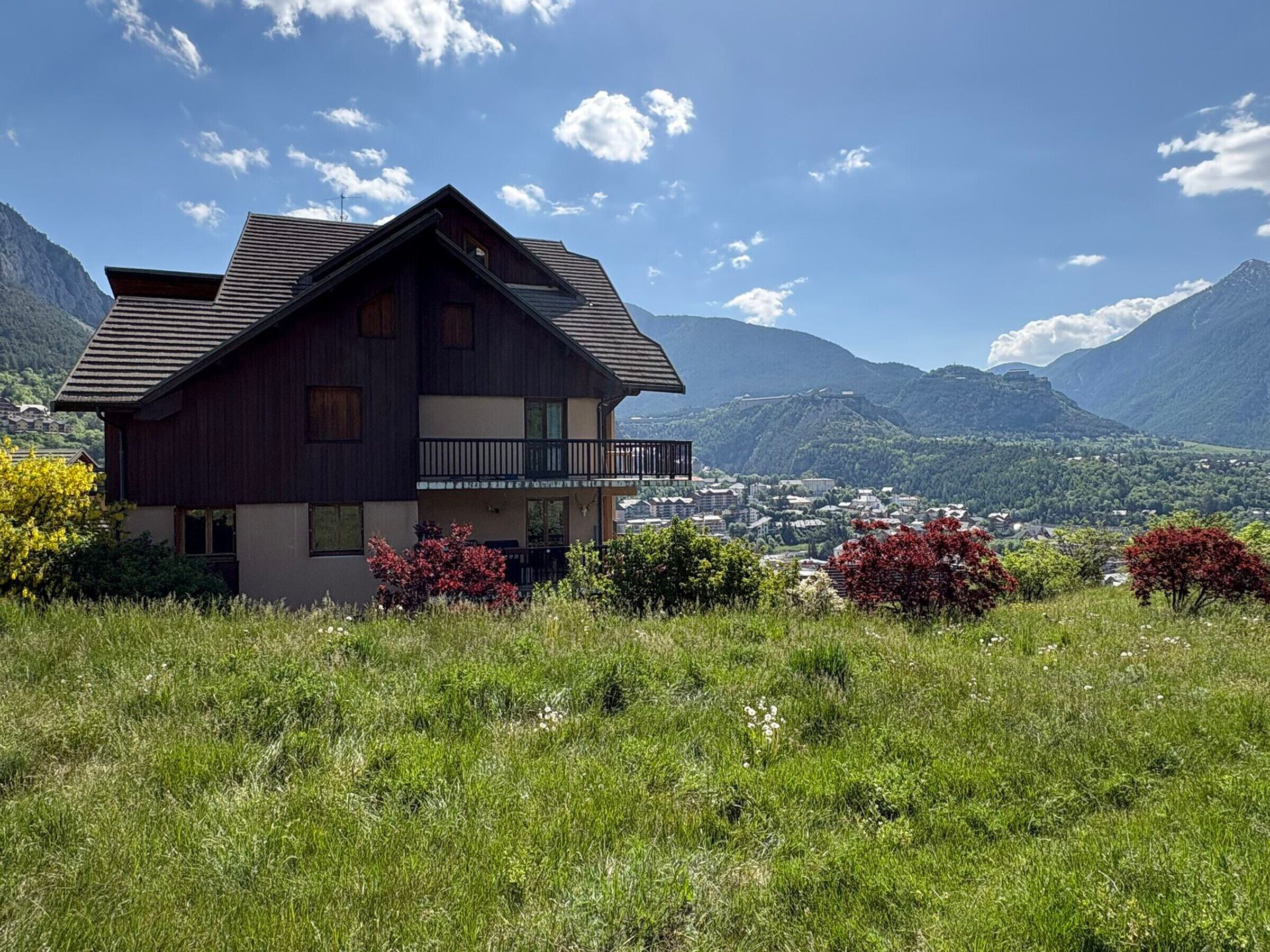 A two-story wooden house with a balcony, surrounded by lush greenery and mountains in the background.