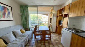 Un espai vital compacte amb sofà, taula de dinar, cadirees, petita cuina i una porta corredissa de vidre que dona a una zona exterior.