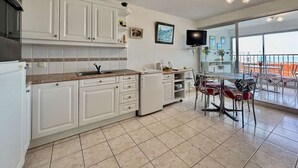 Cozinha com armários brancos, área de jantar com mesa e cadeiras, e varanda com vista.