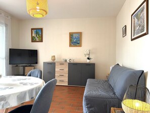 A living room with a sofa, a TV, a dining table with chairs, a cabinet, and two framed pictures on the wall. - Quiet apartment with parking - 4 guests (Le Lavandou)