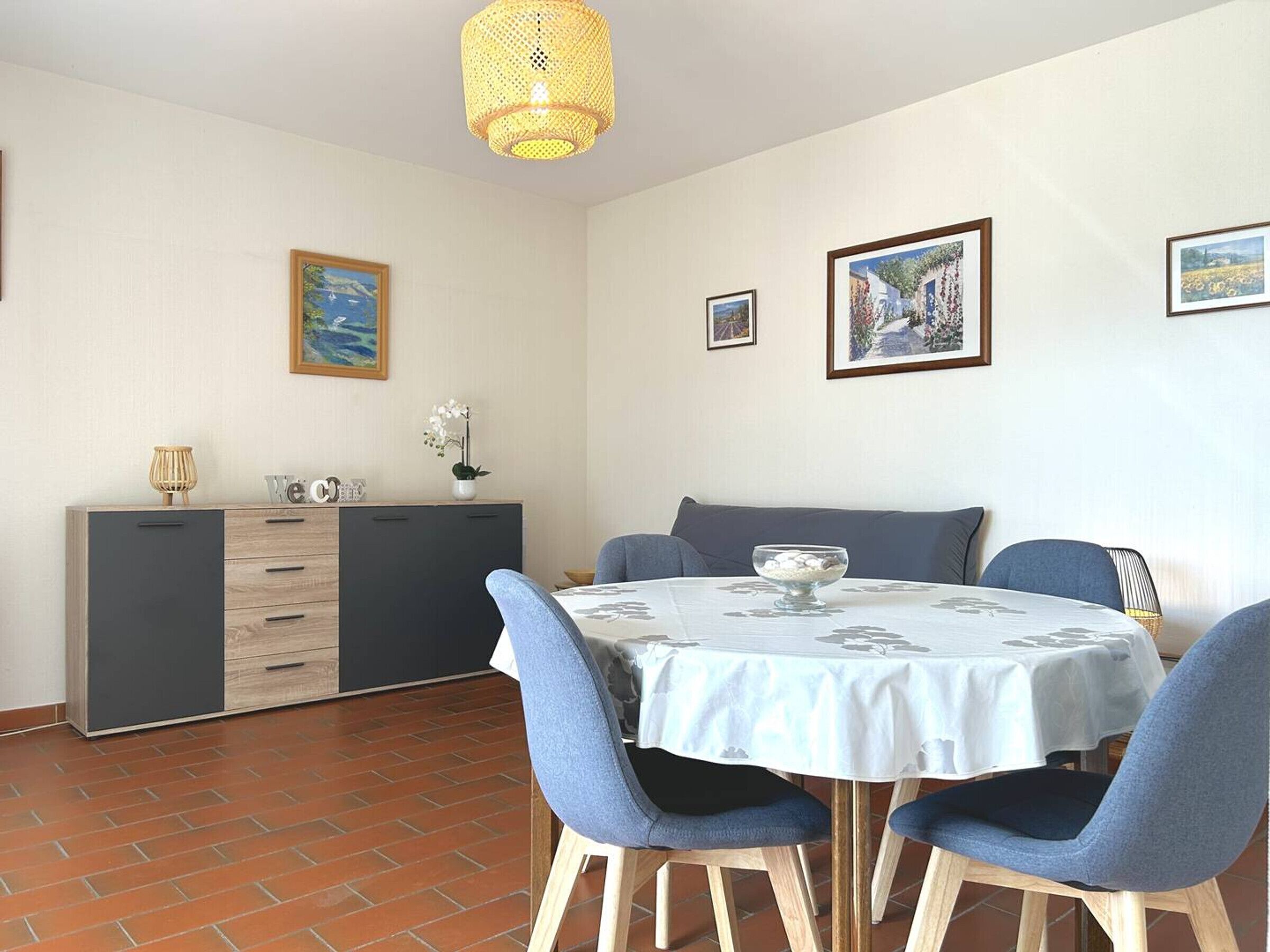 A dining area with a round table, blue chairs, a sofa, a cabinet, and two framed pictures on the wall.