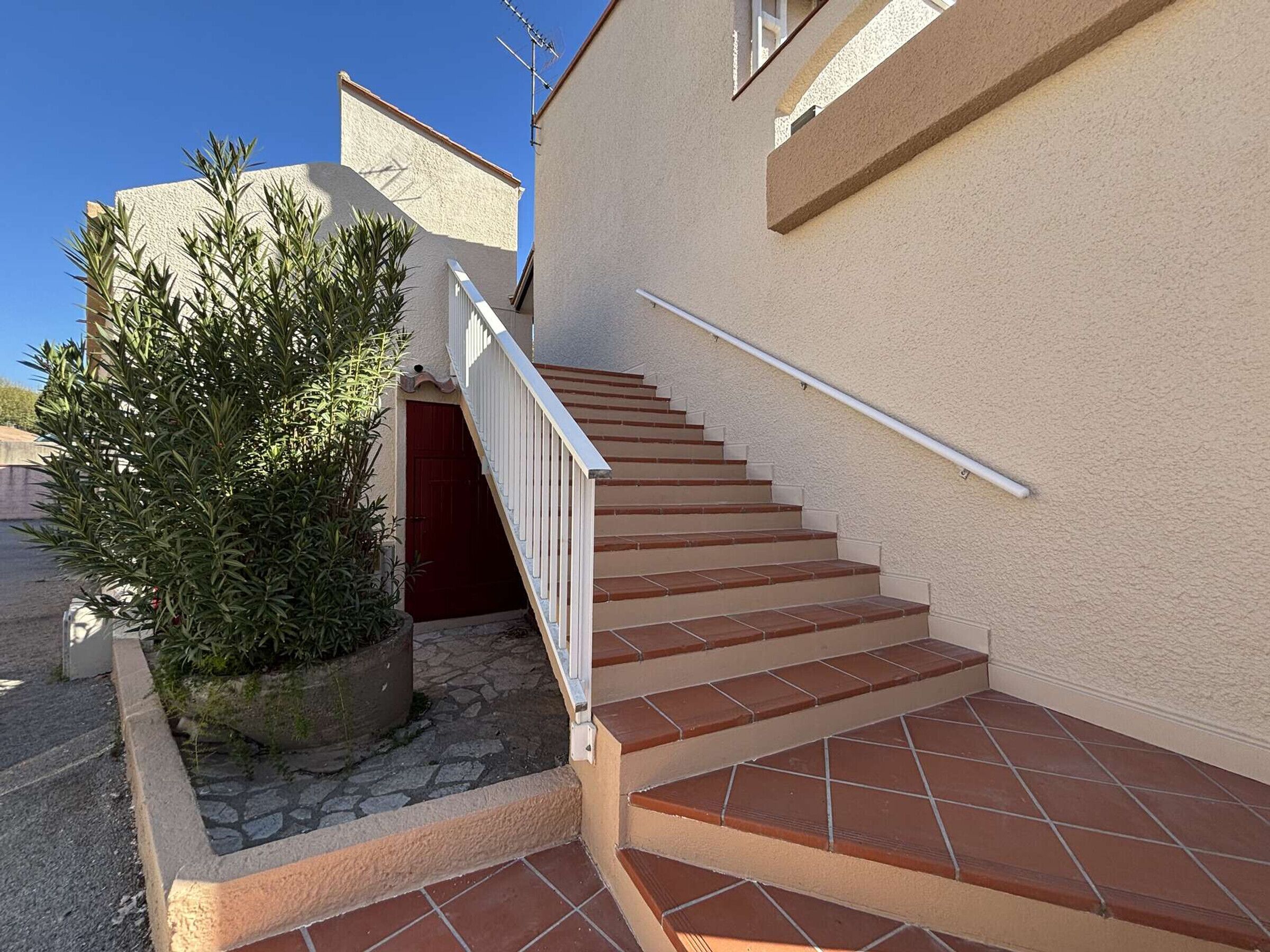 Una escalera conduce a la entrada de un edificio con una puerta roja, rodeada por un pequeño jardín con un arbusto grande.
