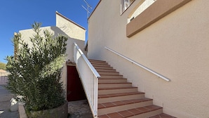Un escalier menant à l’entrée d’un immeuble, avec une porte rouge, le tout entouré d’un petit jardin agrémenté d’un grand arbuste.