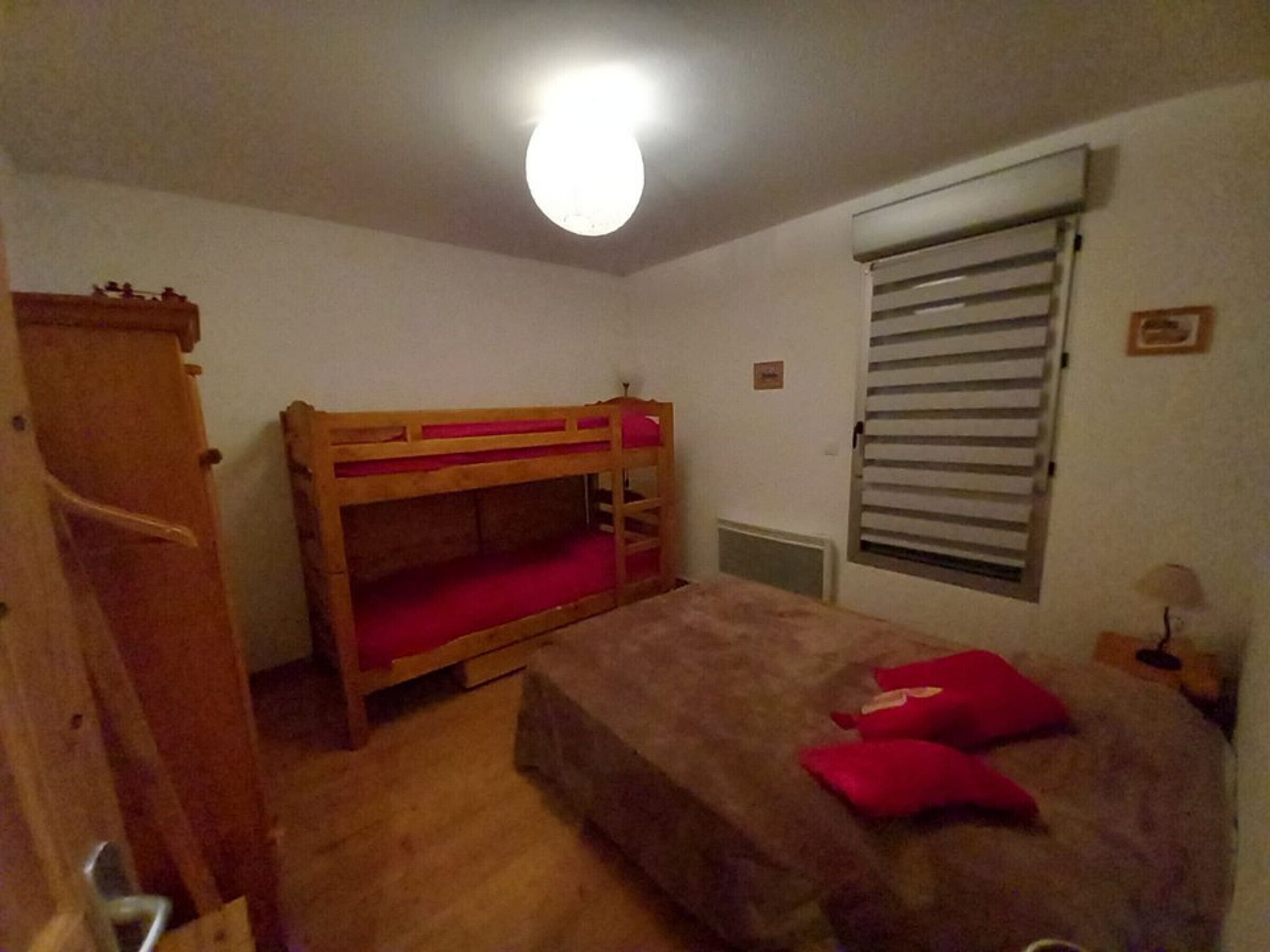 A small, single-room accommodation with a bunk bed, a radiator, a window with blinds, and a bedside table with a lamp.