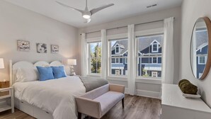 Ein Schlafzimmer mit Bett, Nachttischen, einer Bank, Deckenventilator, großen Fenstern und Blick auf Häuser.