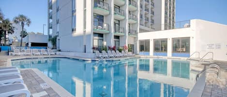 Uma piscina com espreguiçadeiras e um edifício com varandas ao fundo.