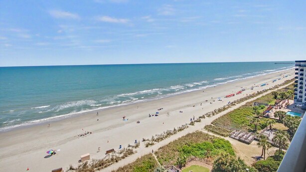 A beach with a long stretch of sand, a pier in the distance, and a building with a pool.