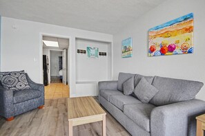A living room with a grey sofa, a blue armchair, and a wooden coffee table. There are framed pictures on the wall and a colorful painting above the sofa. - Ocean 1BR + Beach Access + Dog Friendly (Myrtle Beach)