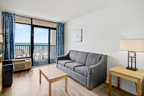 Ein Wohnzimmer mit grauem Sofa, einem Holztisch und Blick auf das Meer durch eine Schiebetür.