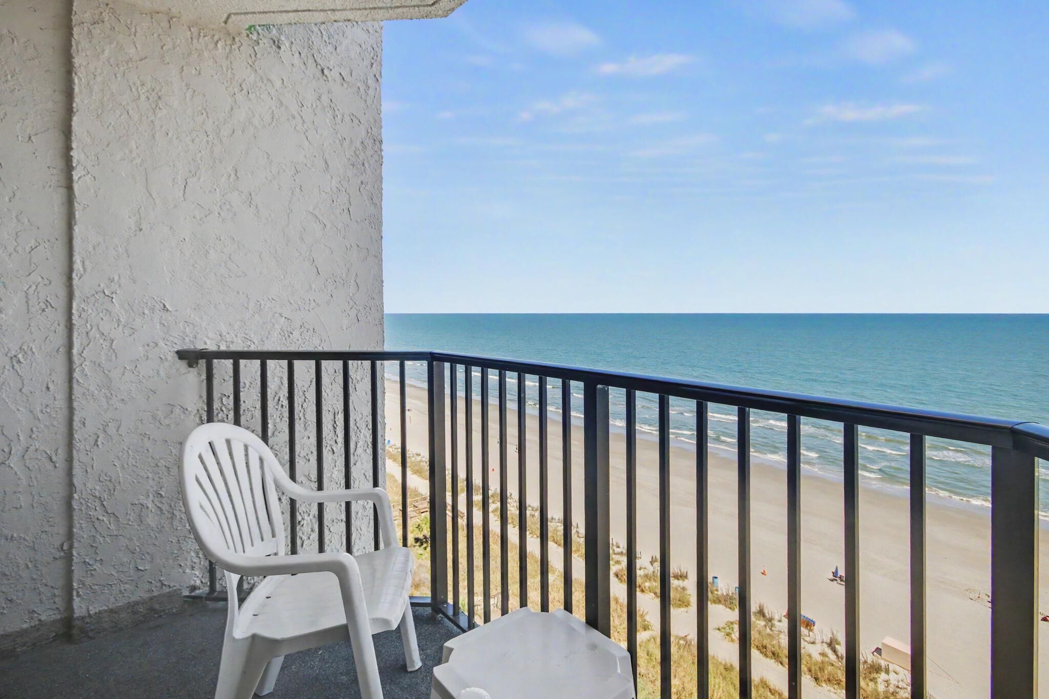 A balcony with a white plastic chair and a white plastic table, overlooking a beach with a clear sky.