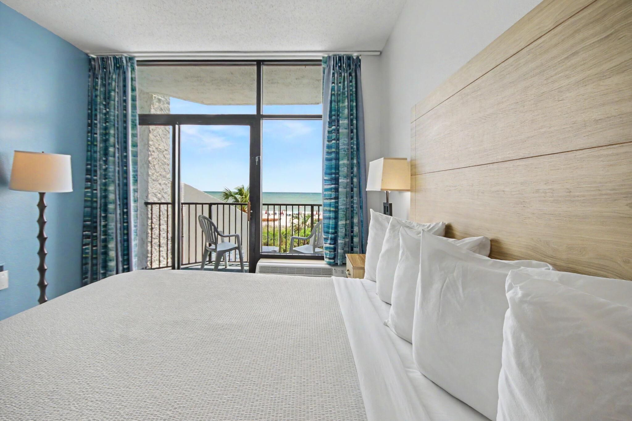 Een hotelkamer met een groot bed, twee bedlampen, uitzicht op het strand en een balkon met stoelen.