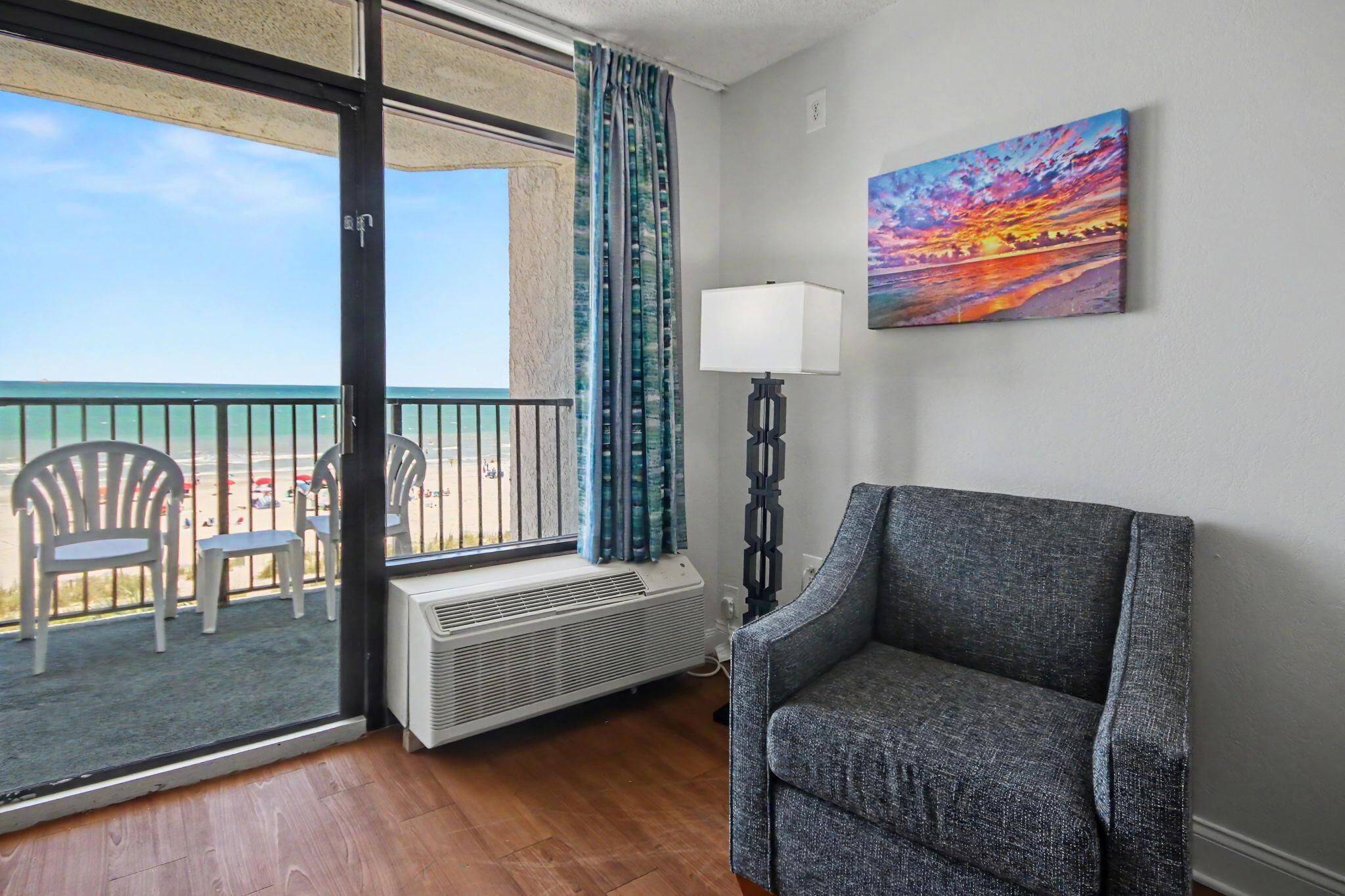 A room with a view of the beach, a chair, a lamp, and a painting on the wall.