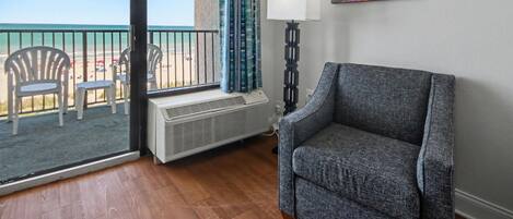 A room with a view of the beach, a chair, a lamp, and a painting on the wall.