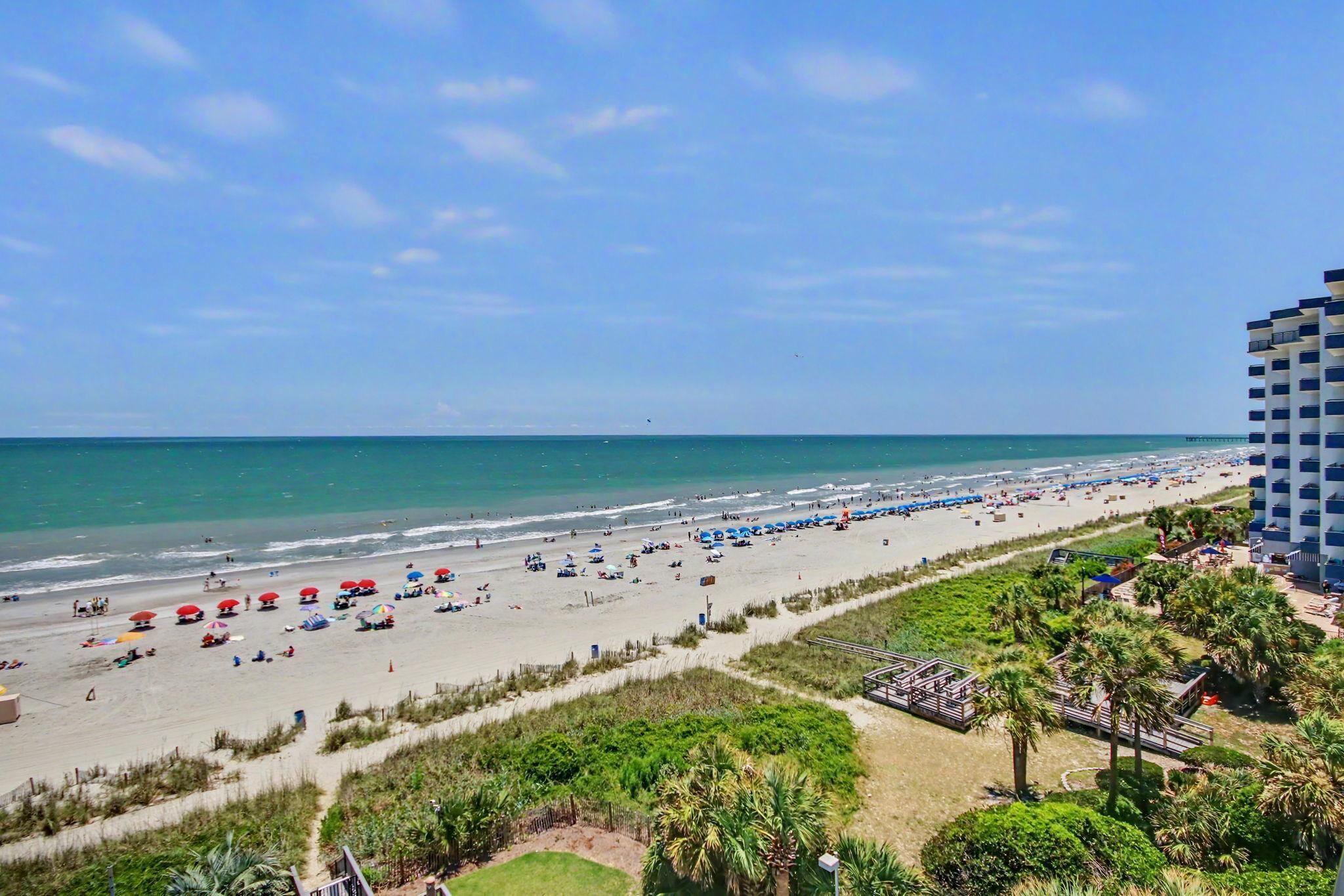 A beach with umbrellas and people, a pier in the distance, and a hotel building on the right.