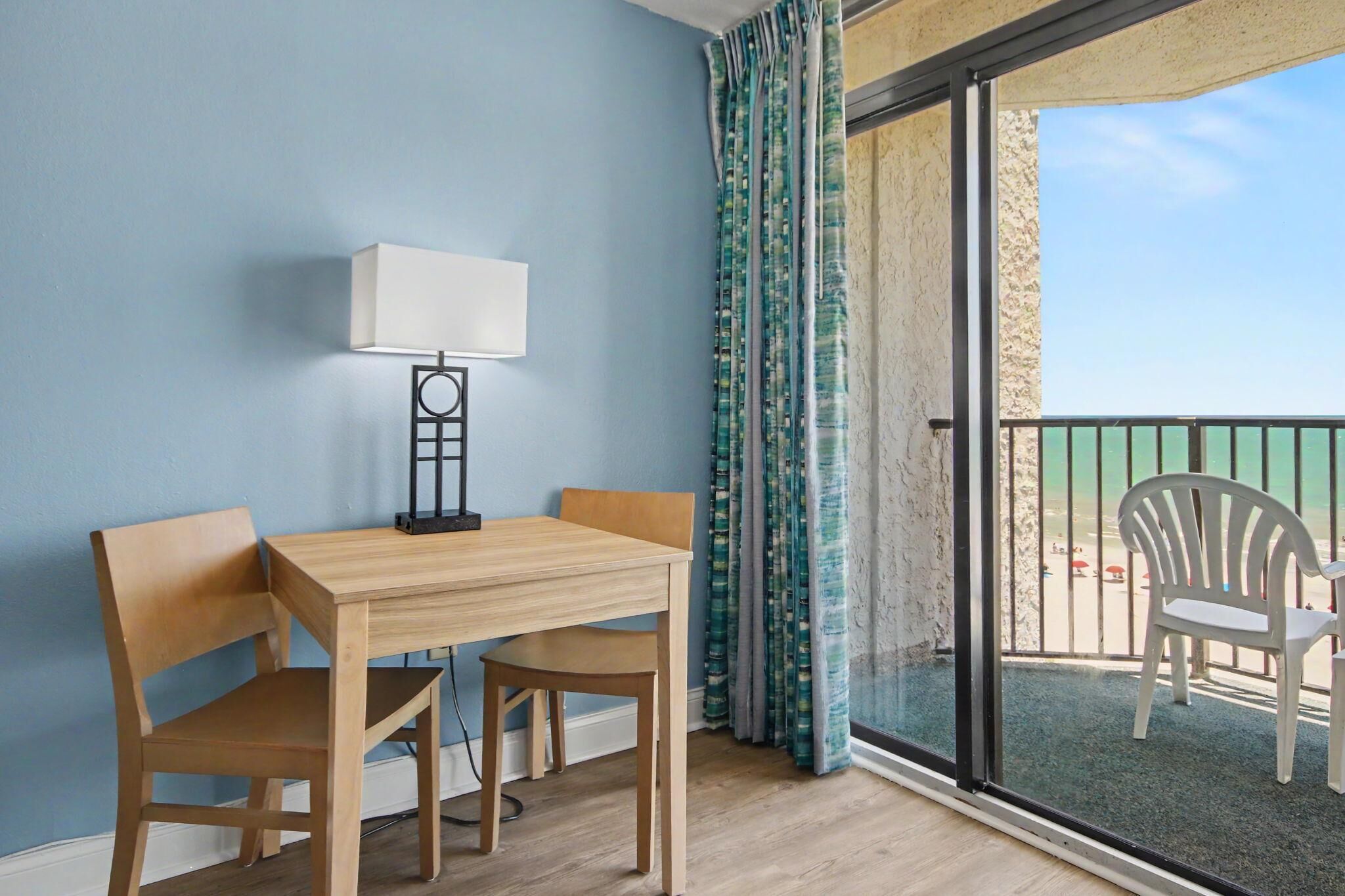 Una habitación con una mesa y sillas de madera, una lámpara de pie y vistas al mar a través de un ventanal.