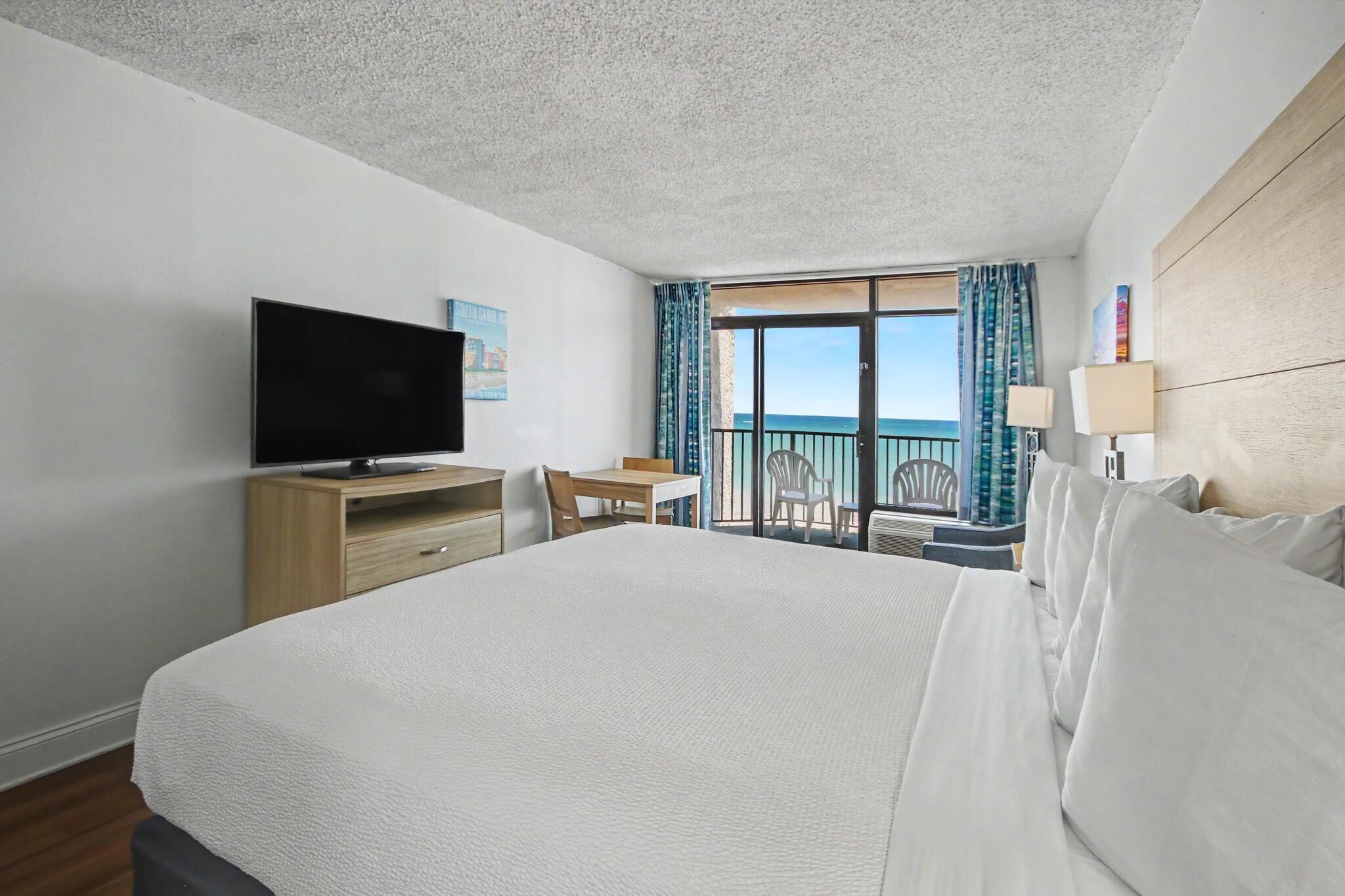 A hotel room with a large bed, a TV on a wooden stand, a balcony with a view of the sea, and a desk with a chair.