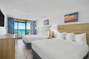 Een hotelkamer met twee bedden, een bureau, een stoel, een balkon met uitzicht op de oceaan en kunst aan de muren.