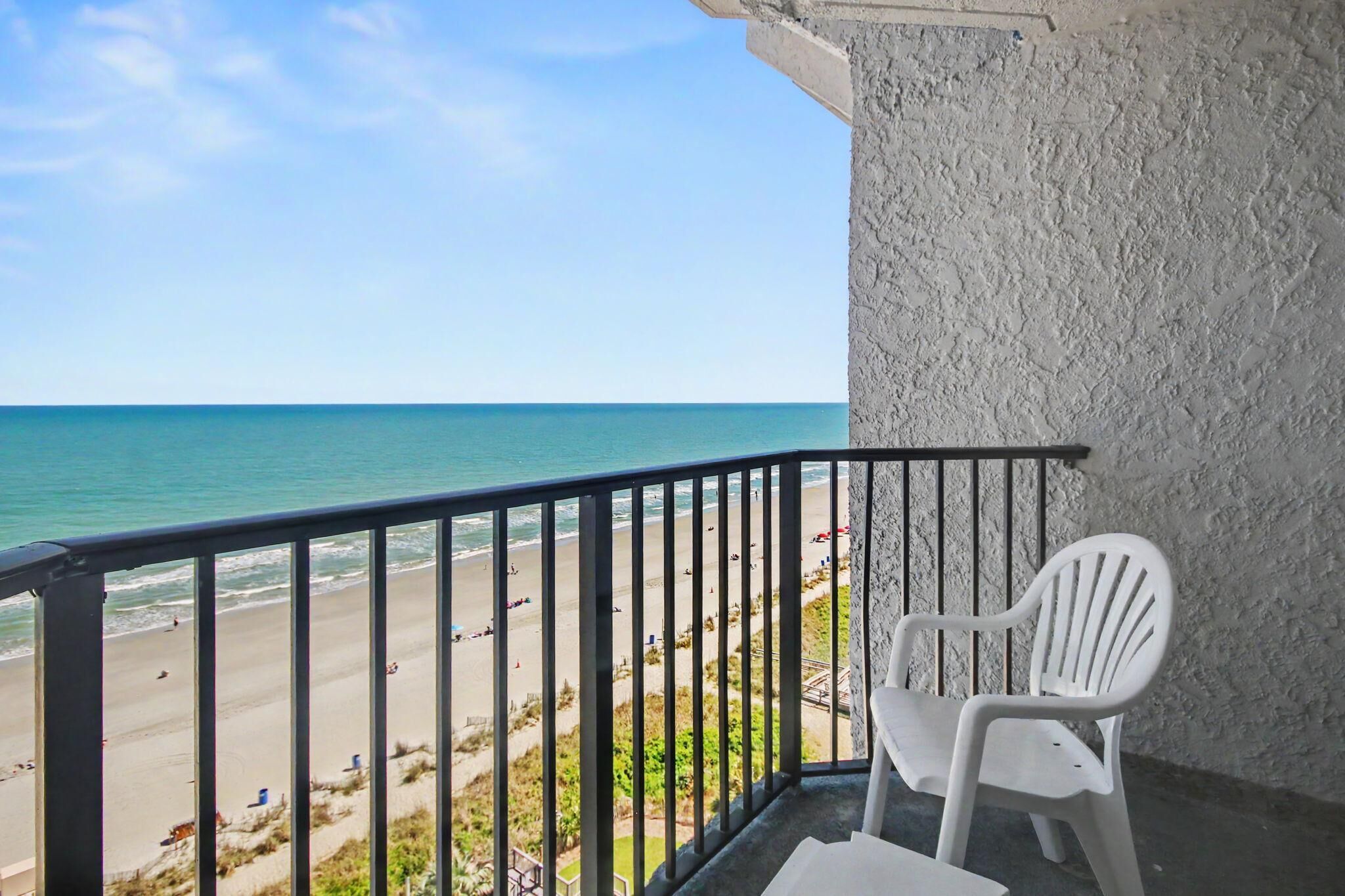 Un balcon avec une chaise en plastique blanche, une vue sur la plage et un ciel dégagé.