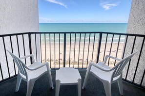 Un balcon avec deux chaises en plastique blanches et une petite table, donnant sur une plage sous un ciel dégagé.