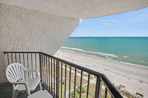 Ein Balkon mit Blick auf einen Strand, ein weißer Plastikstuhl und ein Geländer.