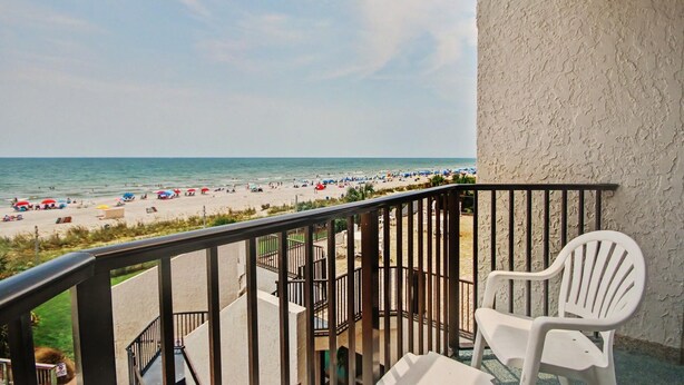 A balcony with a view of a beach, a white plastic chair, and a railing. - Oceanfront Two Queen Room + Beach + Dog Friendly (Myrtle Beach)