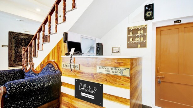 A reception desk with a 'Collection' sign, a 'Reception' sign, a 'Check In / Check Out' sign, a 'Time: 12:00' sign, and a '104' door number. - Deluxe Rest A While (Srinagar)