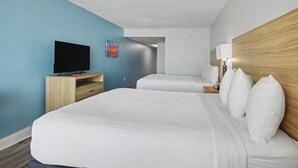A hotel room with a large bed, a TV on a wooden stand, a bedside table with a lamp, and a blue wall. - Oceanfront Two Queen Room + Beach + Dog Friendly (Myrtle Beach)