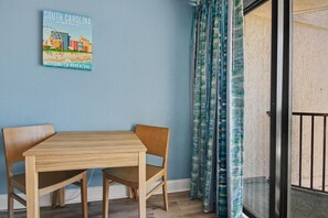 Huoneessa on puinen pöytä ja tuolit, seinälle kiinnitetty kyltti ja ikkunasta avautuva näkymä, jonka edessä on verhot.