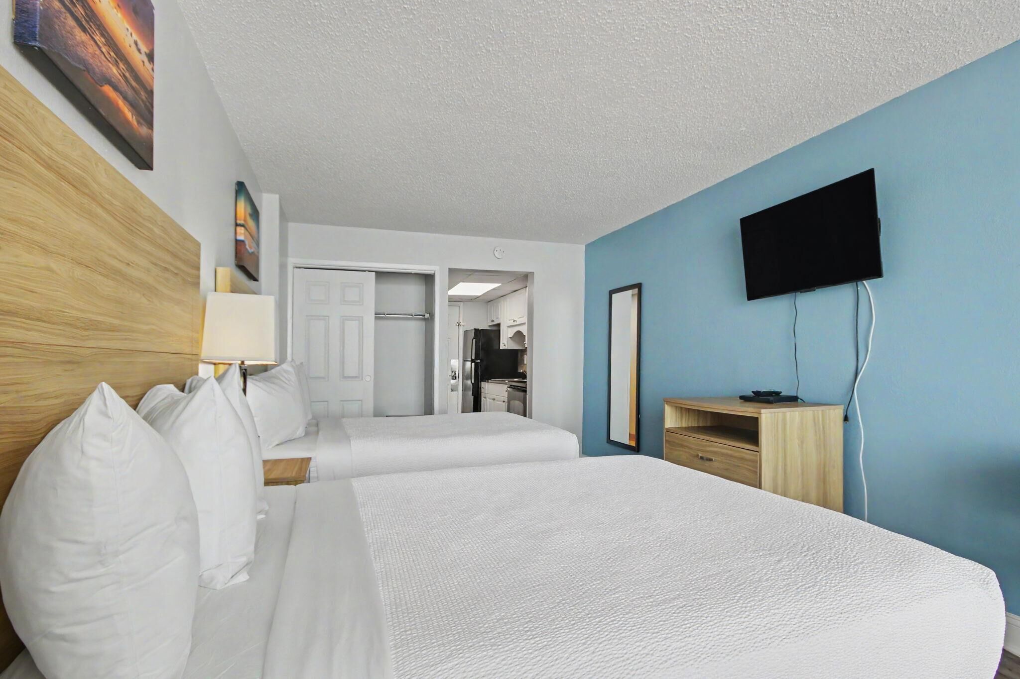 A hotel room with two beds, a television mounted on the wall, and a view into the kitchen area.