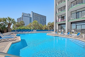 Une piscine avec des chaises longues, un immeuble de grande hauteur en arrière-plan et un ciel bleu dégagé.