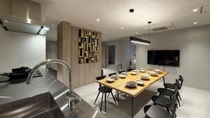 A modern kitchen with a stainless steel range hood, a wooden dining table with chairs, and a flat-screen TV mounted on the wall. - WAKO Ekimae (Hiroshima City)