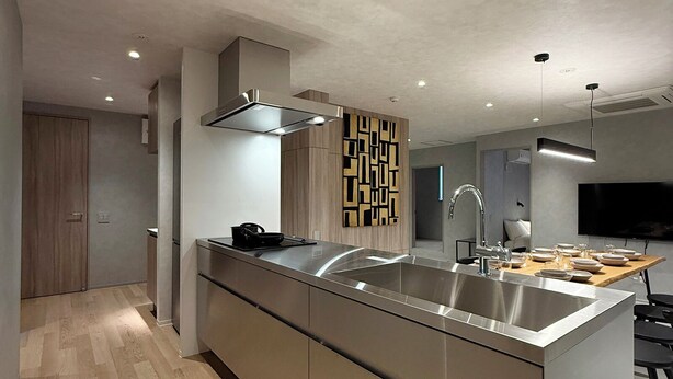 A modern kitchen with a stainless steel island, recessed ceiling lights, and a wooden floor. - WAKO Ekimae (Hiroshima City)