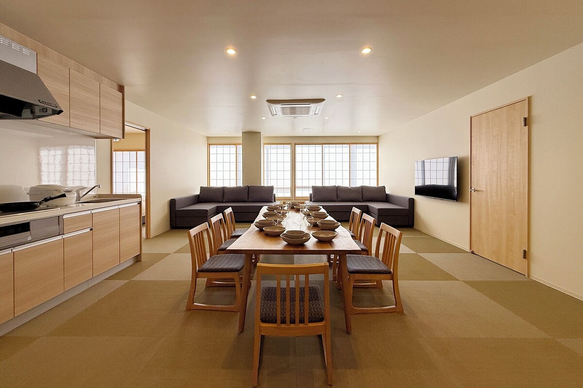 A modern kitchen with a dining table set for six, a sofa, and a TV on the wall.