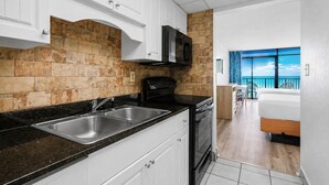 Una cocina con encimera negra, lavaplatos de acero inoxidable y armarios blancos. Hay un horno negro y un microondas sobre la estufa. La cocina tiene un salpicadero de piedra y vistas a un balcón con cama y sillas.