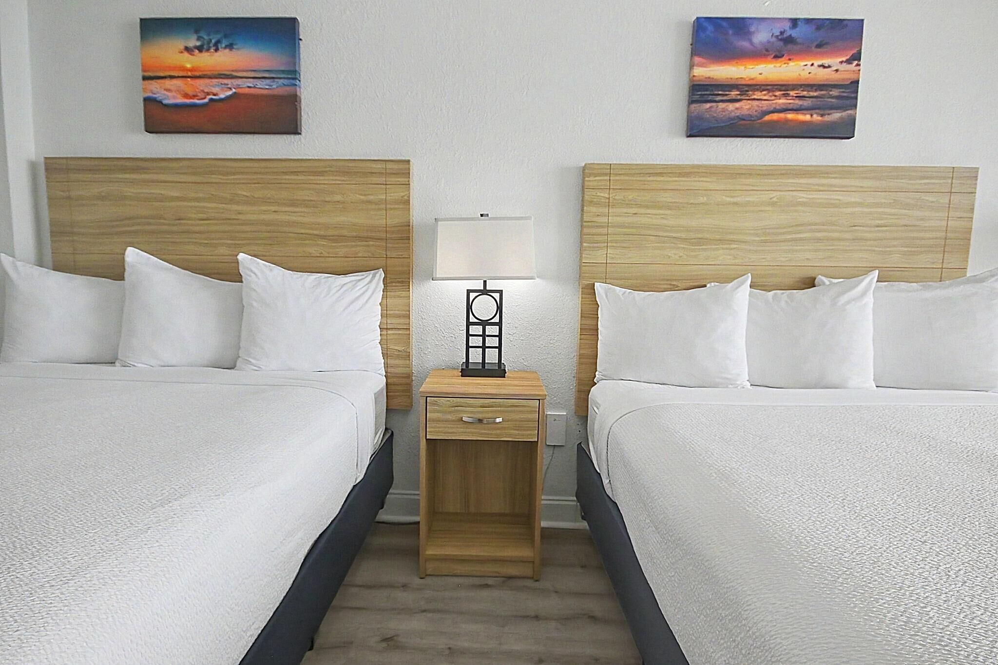 Une chambre d’hôtel avec deux lits, une table de chevet avec une lampe et deux tableaux représentant un coucher de soleil accrochés au mur.