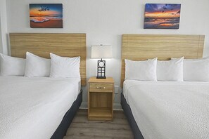 Una habitación de hotel con dos camas, una mesita de noche con una lámpara y dos cuadros con paisajes al atardecer en la pared.