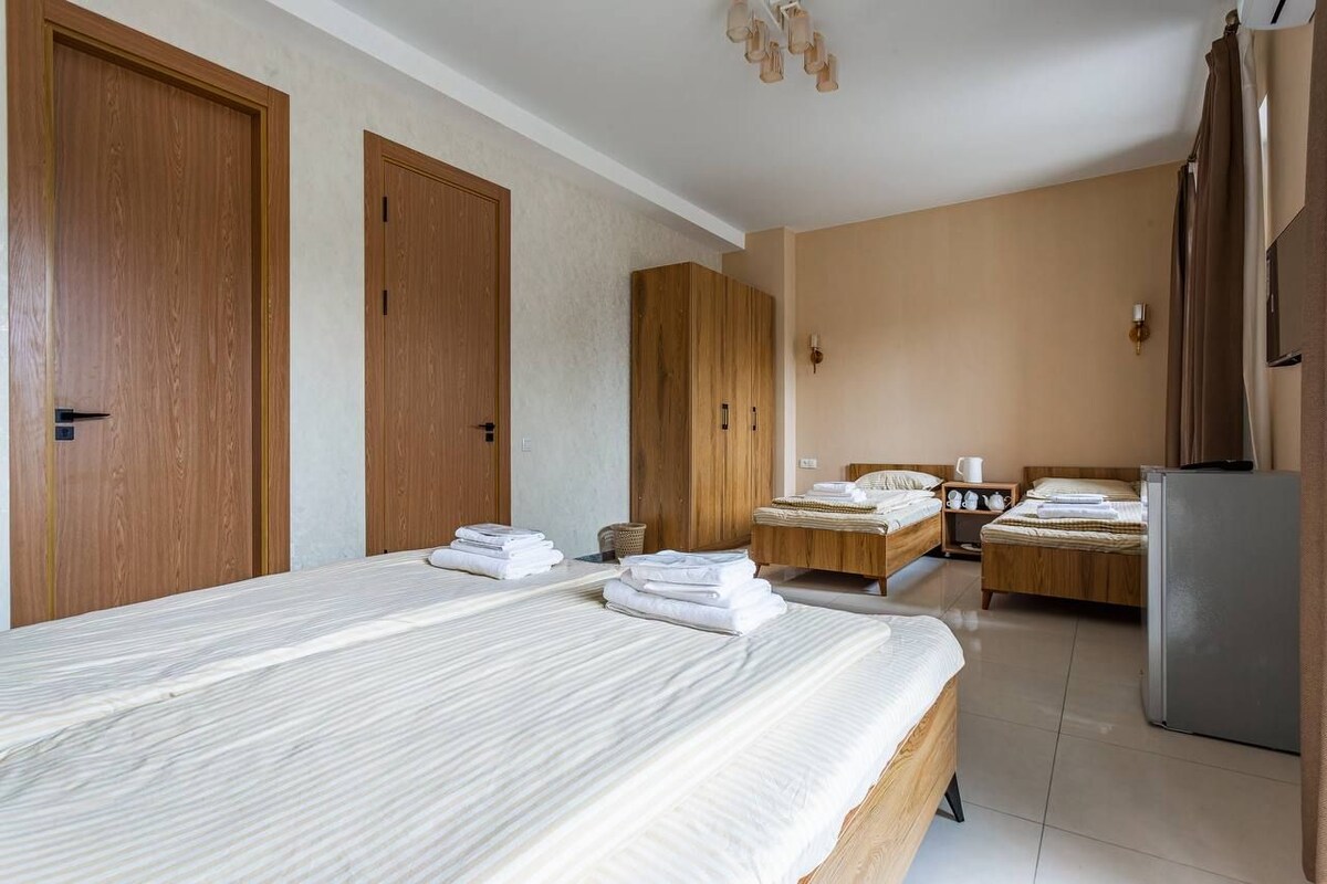 A hotel room with two beds, wooden wardrobes, a refrigerator, and a small table with a cup.